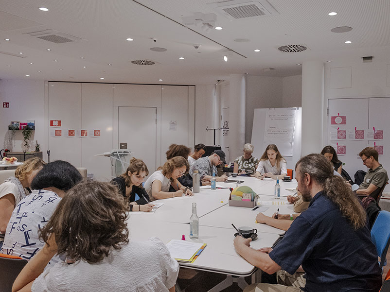 Dieses Bild zeigt viele Personen, die rund um einen großen Arbeitstisch sitzen und sich konzentriert mit dem zur Verfügung gestellten Material zu nachhaltiger und digitaler Transformation auseinandersetzen. 