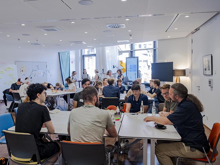 Dieses Bild zeigt mehrere Workshop-Gruppen, die um Tische verteilt sitzen und arbeiten.