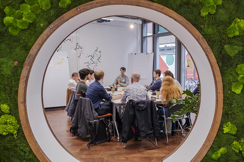 Ein begrünter Torbogen gibt den Blick frei auf einen Tisch, an dem eine Workshop-Gruppe gemeinsam arbeitet.