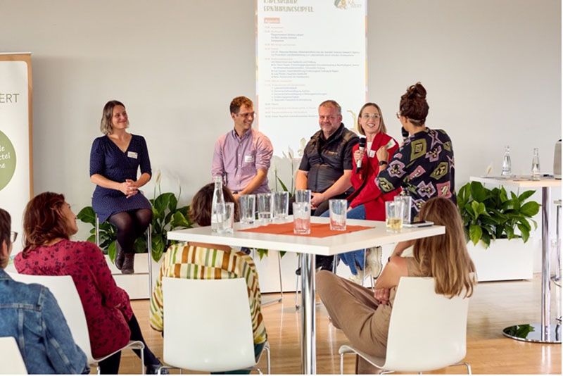 Podiumsdiskussion mit mehreren Teilnehmern und Zuhörern in einem modernen Raum.