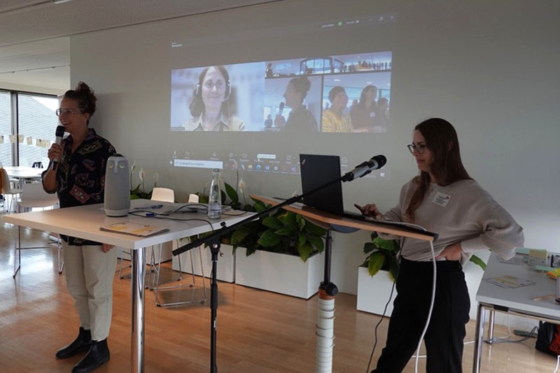 Zwei Personen halten eine hybride Präsentation vor Publikum und Video-Teilnehmern im modernen Raum.
