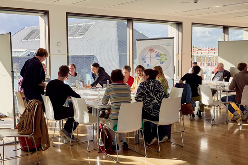 Teilnehmer eines Workshops sitzen an Tischen in hellem Seminarraum mit großem Fenster.