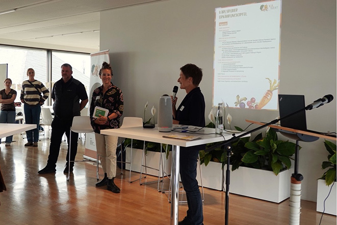 Bettina Lisbach hält Vortrag mit Mikrofon vor Publikum, Wandpräsentation im Hintergrund sichtbar.