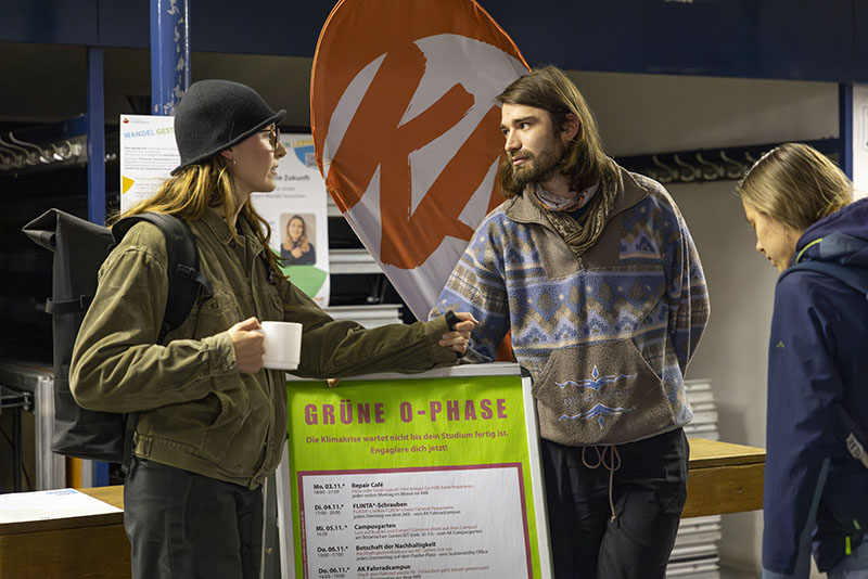 Personen stehen an einem Aufsteller der Grünen O-Phase, dahinter eine Flagge vom Karlsruher Transformationszentrum.