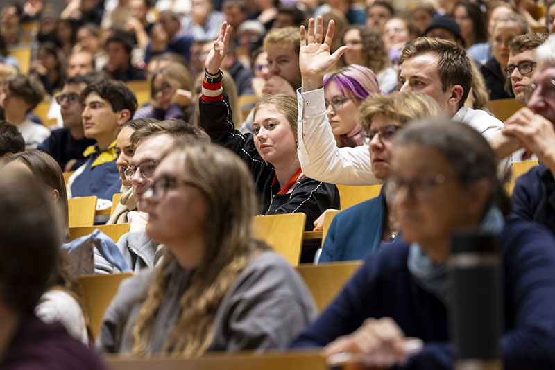 Hörsaal voller Studierender, einige melden sich mit erhobenen Händen.