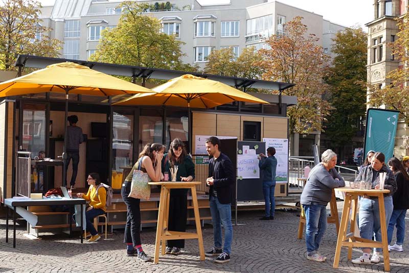 Menschen unter Sonnenschirmen an Stehtischen auf einem Platz in Karlsruhe bei sonnigem Herbstwetter.