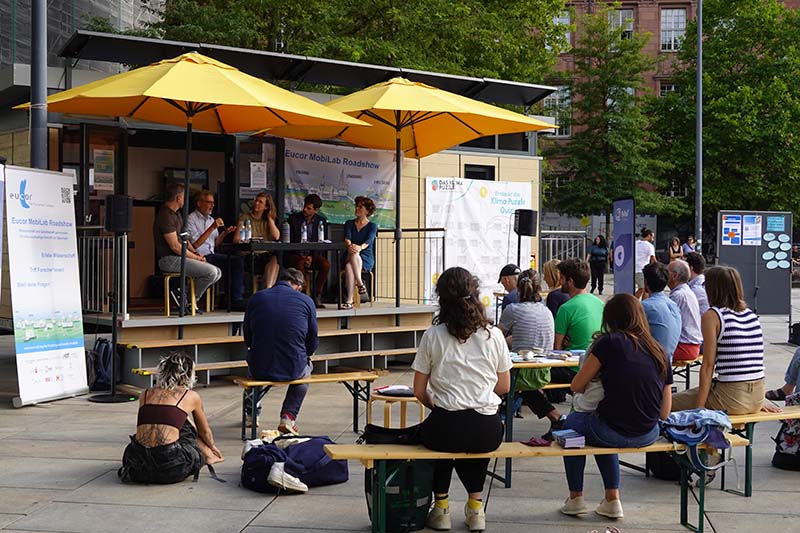 Das MobiLab im Freien mit Publikum unter gelben Sonnenschirmen bei der Roadshow in Freiburg.