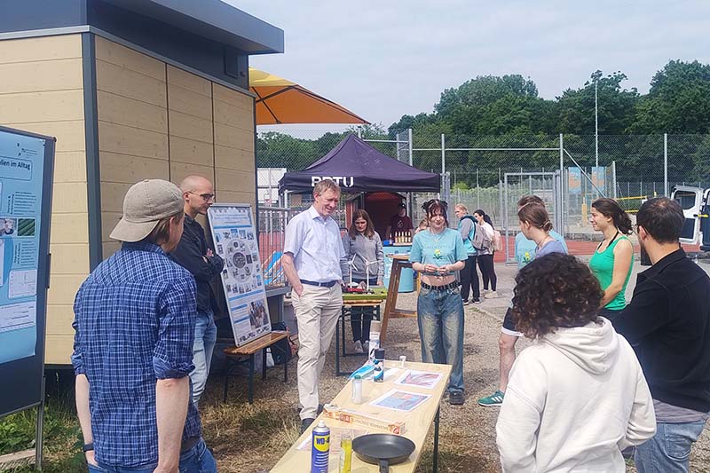 Outdoor-Informationsstand mit mehreren Personen vor einem Pavillon und Plakaten bei Sonnenschein.