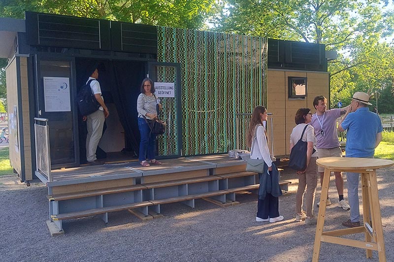 Menschen vor und in einem mobilen Ausstellungscontainer im Freien bei sonnigem Wetter.