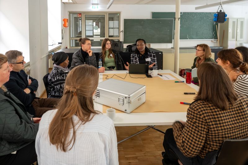 Gruppe von Menschen sitzt um einen Tisch in einem Seminarraum, Moderator mit Laptop.