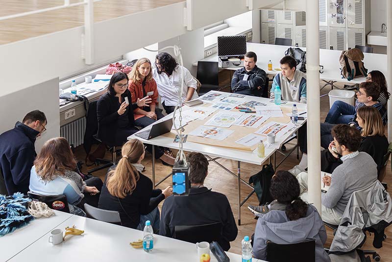 Gruppe junger Menschen sitzt um einen Tisch mit Unterlagen, Laptops und Druckmaterialien in einem B&uuml;roraum.