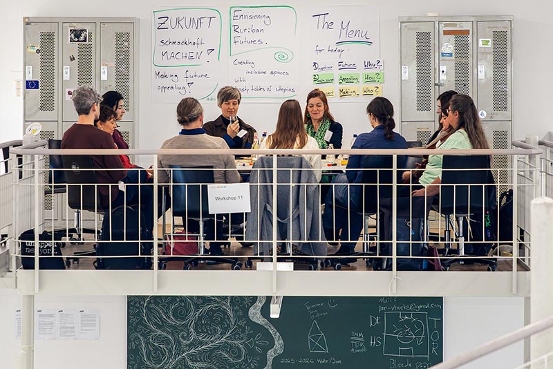 Gruppe von Forschenden sitzt an einem Tisch in einer Aula, Whiteboard mit Notizen im Hintergrund.