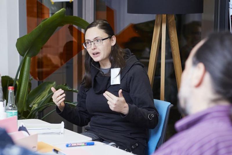 Frau erklärt sitzend an einem Tisch in einem modernen Büro mit Pflanzen und Lampen.