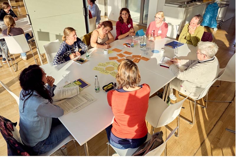 Gruppe Menschen sitzt um einen Tisch bei einem Workshop. Kärtchen, Stifte und Unterlagen sind auf dem Tisch verteilt.