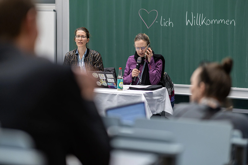 Zwei Frauen sprechen bei einer Veranstaltung vor einer grünen Tafel mit der Aufschrift „Willkommen“.