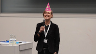 Lachende Frau mit Mikrofon und rosa Partyhut bei einer Veranstaltung.