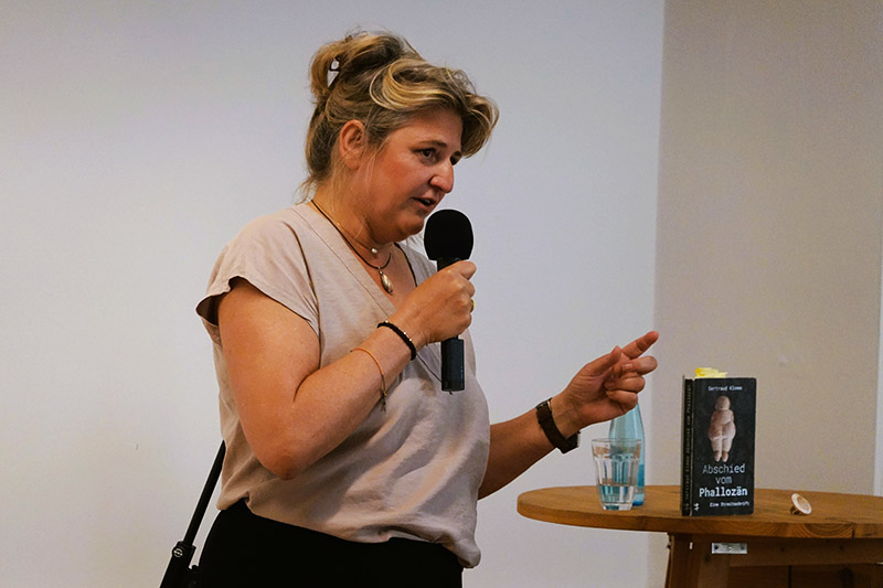 Frau spricht mit Mikrofon, auf einem Stehtisch ist ein Buch mit dem Titel „Abschied vom Phallozän“.