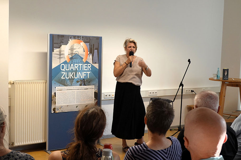 Frau spricht mit Mikrofon vor Publikum neben einem Roll-up-Banner „Quartier Zukunft“.