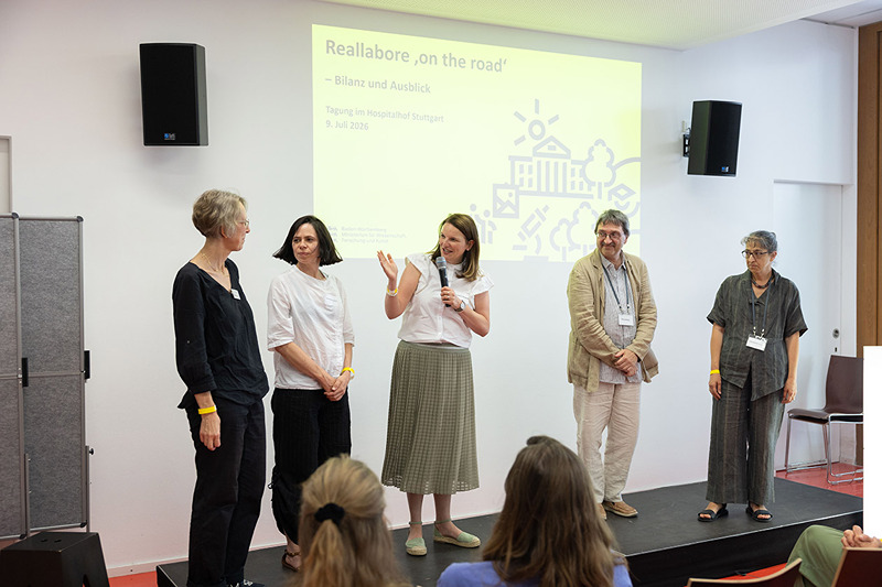 Personen auf einer Bühne begrüßen das Publikum zur Tagung "On the Road" in Stuttgart.