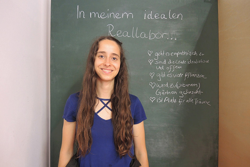 Eine Person steht vor einer Tafel mit Text darauf und lächelt in die Kamera.