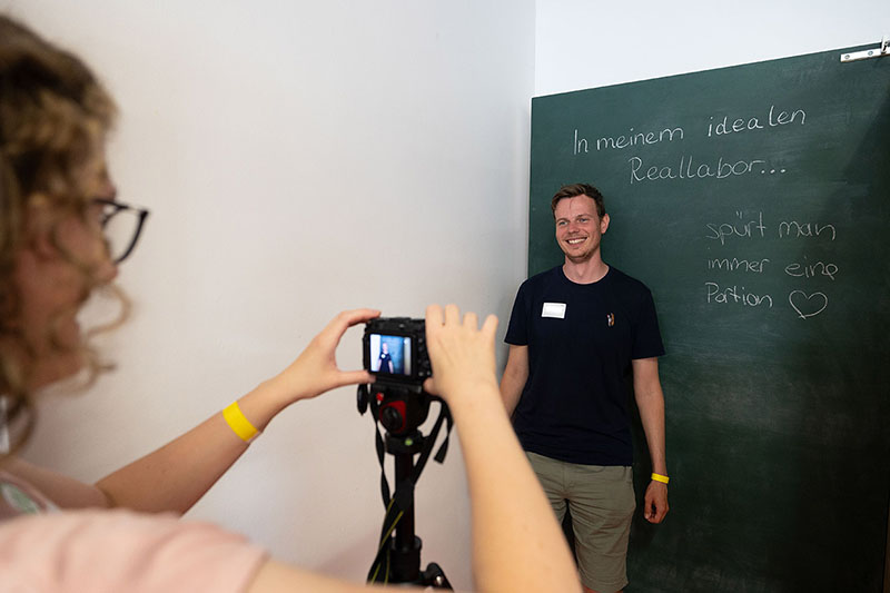Eine Person steht in einer Fotobox mit einer Kreidetafel und einem Spruch darauf und wird gerade fotografiert.