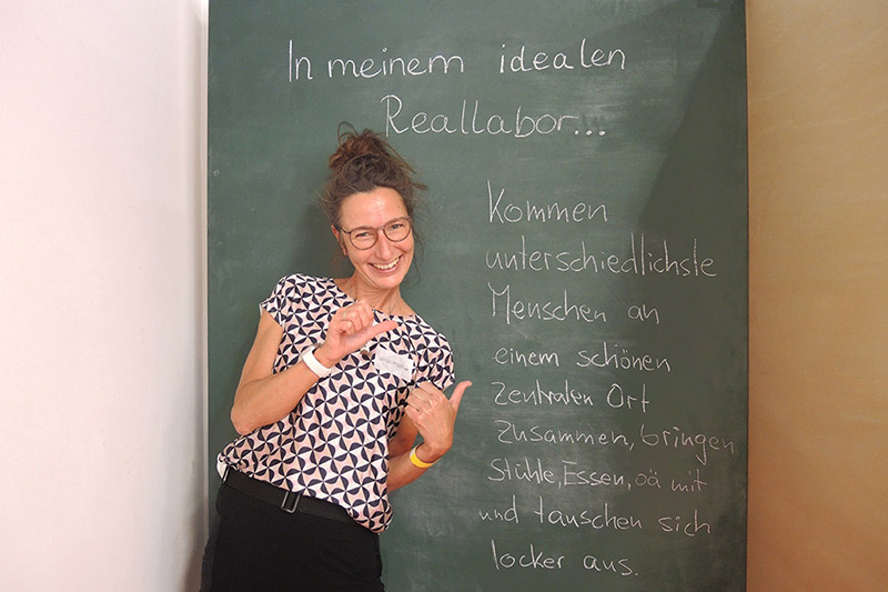Eine Person steht vor einer Tafel mit dem Text darauf "in meinem idealen Reallabor" und lächelt in die Kamera.