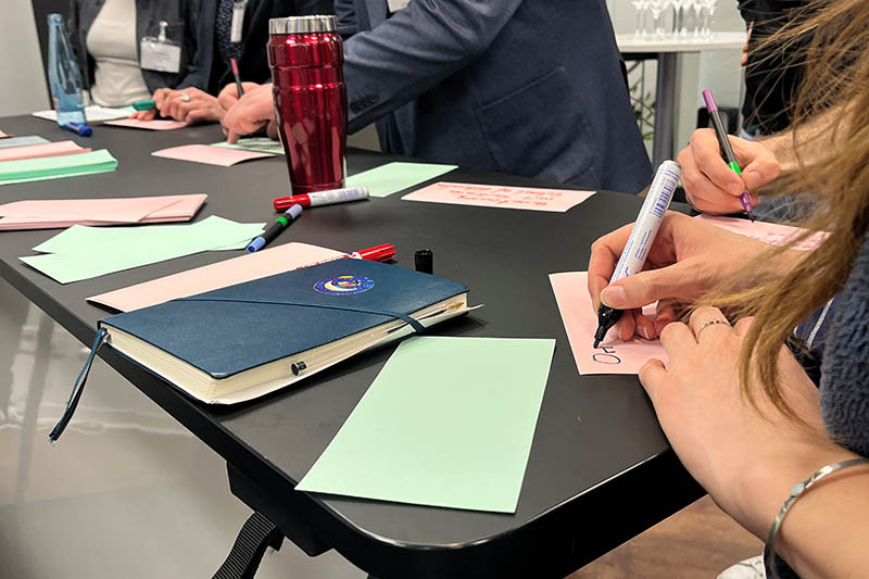 Personen schreiben auf farbige Zettel an einem Tisch; Notizhefte, Stifte und Marker liegen verstreut.