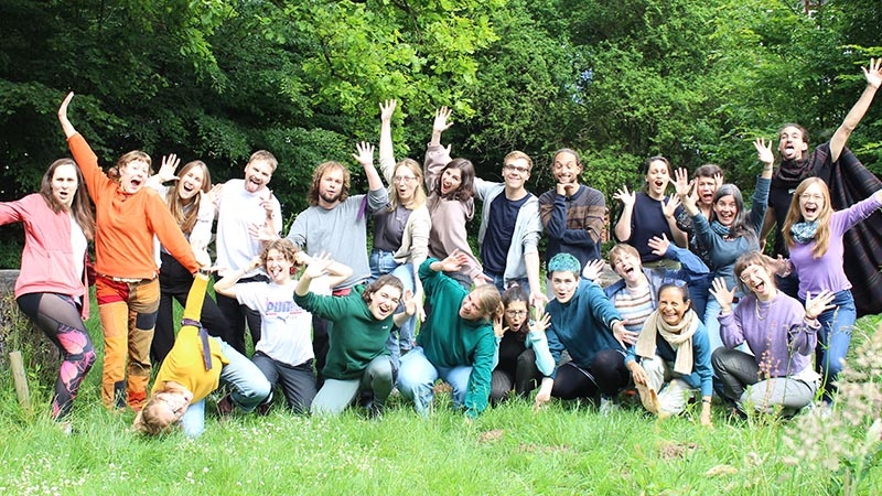Gruppe junger Menschen posiert lachend und gestikulierend in einer grünen Naturlandschaft.