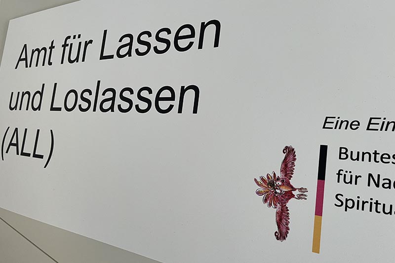 Schild mit der Aufschrift „Amt für Lassen und Loslassen (ALL)“ und einem illustrierten bunten Vogel.