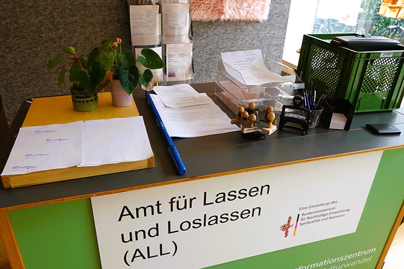 Schreibtisch mit dem Schild „Amt für Lassen und Loslassen (ALL)“ sowie Büromaterial und Pflanzen.