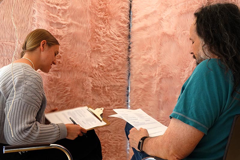 Zwei Personen mit Klemmbrett führen Gespräch; dahinter ein Wandteppich in rosa.