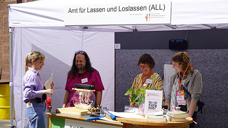 Vier Personen an einem Infostand des „Amts f&uuml;r Lassen und Loslassen (ALL)“.
