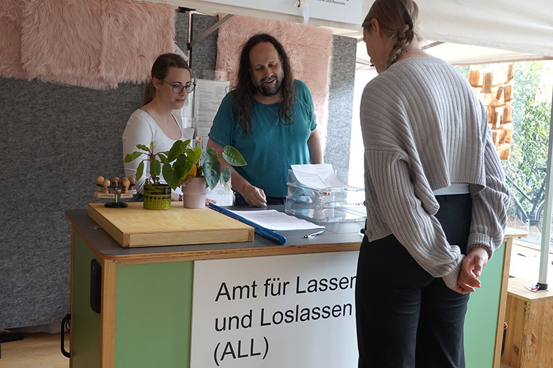 Zwei Personen an einem Stand mit dem Schild „Amt für Lassen und Loslassen (ALL)“ beraten eine dritte Person.