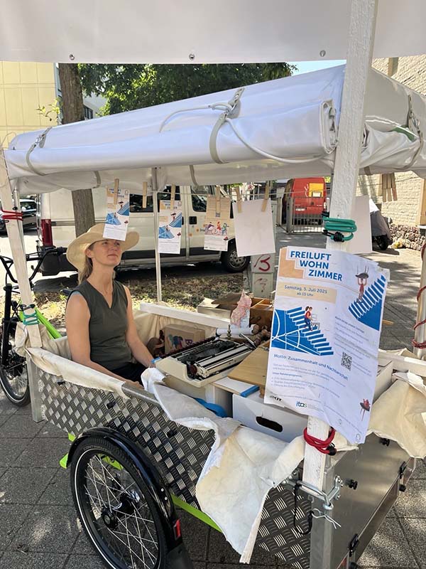 Person sitzt hinten in einem Lastenrad, davor hängen Flyer zum Freiluftwohnzimmer.