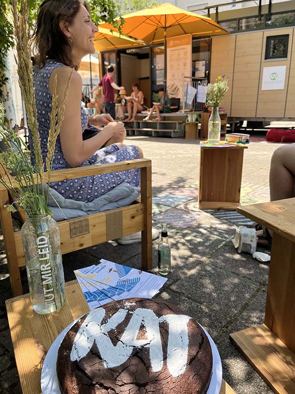 Im Vordergrund ein Kuchen mit KAT-Logo, dahinter fröhliche Personen im Freien. 