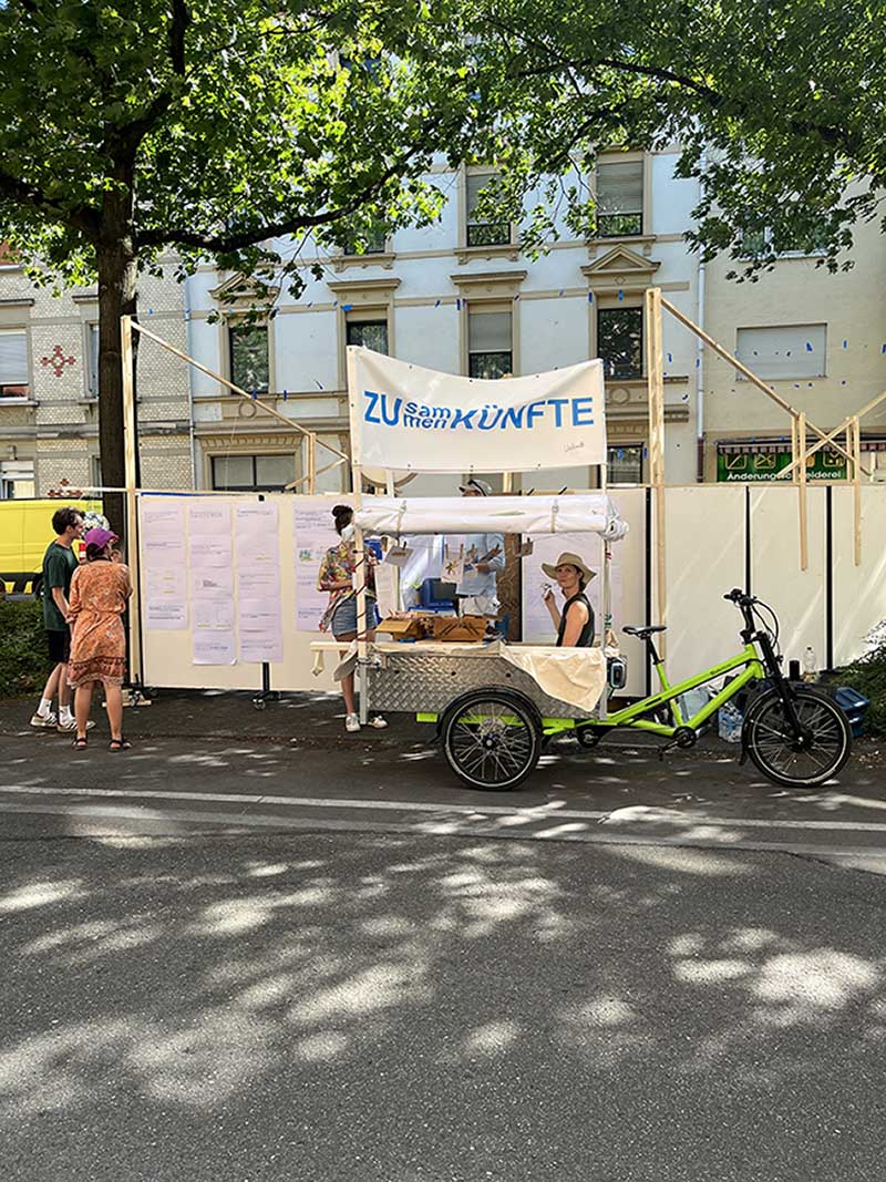 Ein Lastenrad mit einer Person im Wagen steht im Freien vor einem Freiluftwohnzimmer. 