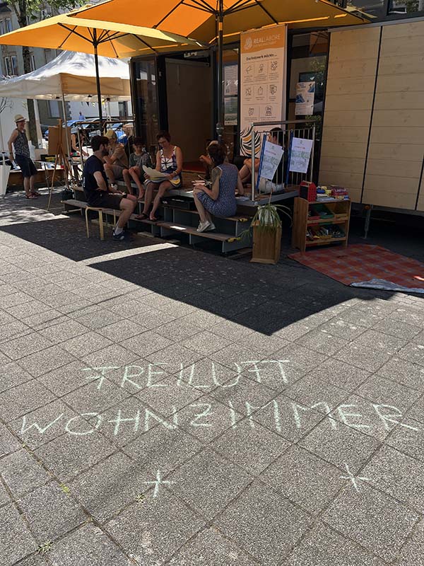 Personen sitzen im Freien vor dem MobiLab, davor der Schriftzug „Freiluftwohnzimmer“ mit Kreide auf den Boden geschrieben. 