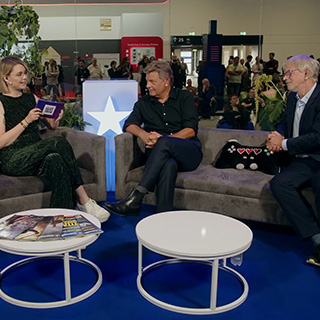 Drei Personen im Gespräch in einer Messehalle, links die Moderatorin mit Tablet, daneben sitzen zwei Männer.
